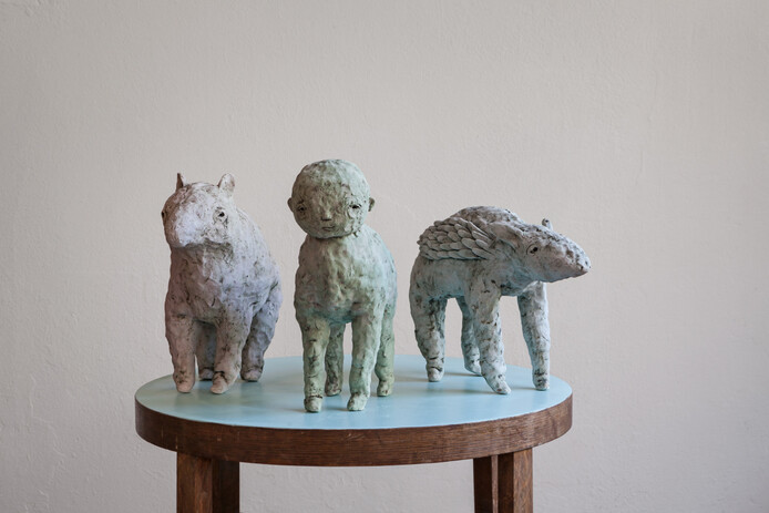 Drei nebeneinander stehende unterschiedliche Tierfiguren aus Keramik auf einer runden Oberfläche. Die Figur in der Mitte trägt einen menschlichen Kopf. 
