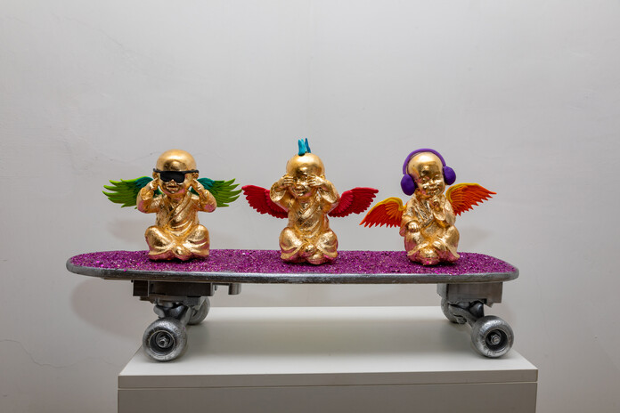 drei vergoldete Figuren mit Flügeln auf einem pinken Glitzerskateboard